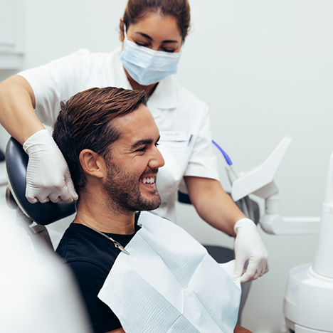 Man sitting in dental chair smiling while dentist adjusts protective napkin