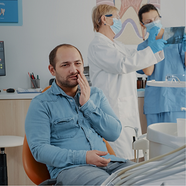 Man in dental chair rubbing jaw in pain while dentists work in background